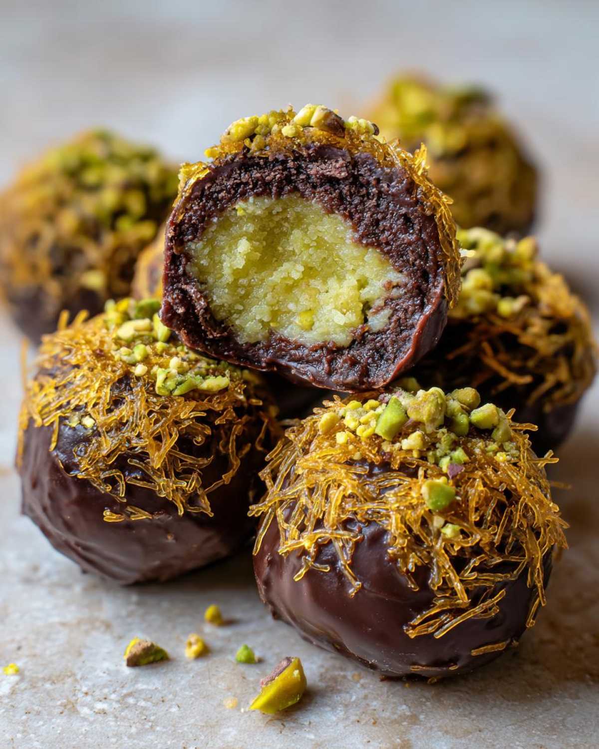 Close-up of dubai chocolate balls, one cut open revealing a yellow filling, coated in chocolate, pistachios, and shredded pastry.