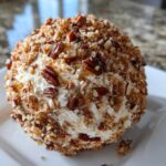 A close-up of a perfectly formed cheese ball recipe coated entirely in chopped pecans and served on a white square plate.
