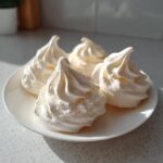 Four perfectly piped, white meringue cookies sitting on a small white plate in bright sunlight.