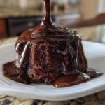 Thick, glossy chocolate syrup being poured over a small, dark chocolate cake slice on a white plate.