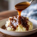 A spoonful of rich homemade brown gravy is being poured over a mound of fluffy mashed potatoes in a bowl.