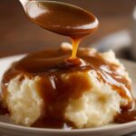 A spoonful of rich, homemade brown gravy being poured over a mound of fluffy mashed potatoes.