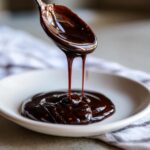 A spoonful of thick, glossy homemade chocolate syrup is being poured onto a white plate.