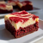 A single square of red velvet cheesecake brownies showing a dense red base and a creamy, swirled cheesecake topping.