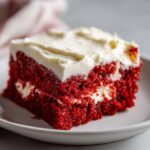 Close-up of a moist slice of red velvet poke cake topped with thick white cream cheese frosting on a white plate.