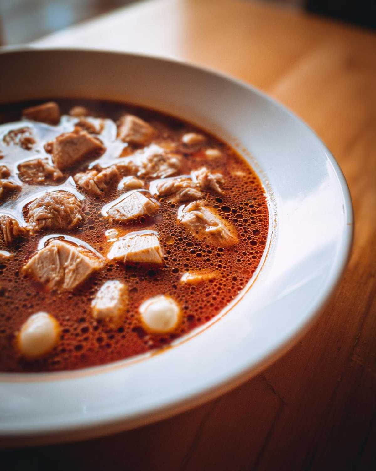 Close-up of rich, red broth chicken pozole with chunks of chicken and hominy in a white bowl.