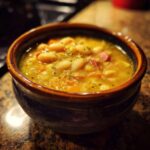 Close-up of a rustic bowl filled with thick, yellow 15 bean soup, featuring white beans and pieces of ham.