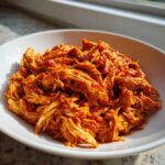 Close-up of flavorful shredded chicken tinga coated in rich, red sauce in a white bowl.