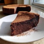 A decadent slice of flourless chocolate torte dusted heavily with cocoa powder, served on a white plate.