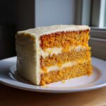 A tall, moist slice of layered Creamsicle Cake with orange-hued layers and white cream cheese frosting.