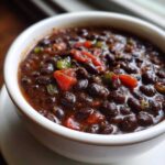 Close-up of a white bowl filled with rich, dark black bean chili, featuring visible chunks of tomato and green pepper.