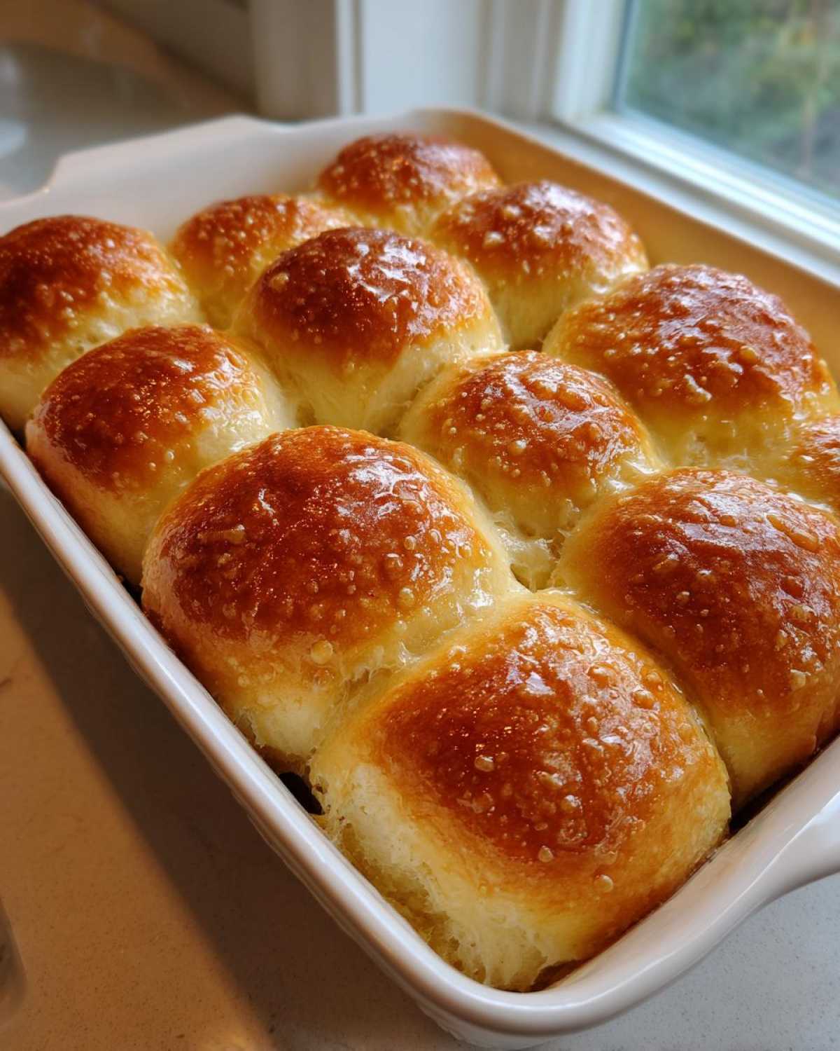 A close-up of golden brown, glossy sourdough discard rolls baked together in a white ceramic dish.
