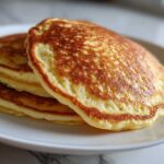 A close-up stack of three golden-brown almond flour pancakes on a white plate.