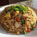 A mound of freshly made chicken fried rice featuring visible pieces of chicken, peas, carrots, and egg, topped with bright green chopped scallions.