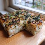 A square portion of baked sushi bake, showing the creamy interior and crispy, seasoned topping of furikake and sesame seeds.