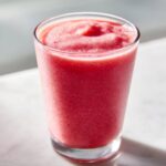 Close-up of a vibrant, icy watermelon smoothie served in a clear glass on a light surface.