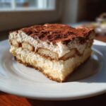 A perfect slice of tiramisu cheesecake layered with cream and ladyfingers, dusted heavily with cocoa powder.