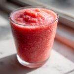 Close-up of a thick, bright red watermelon smoothie served in a clear glass, sitting in sunlight.