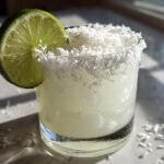 A frosty white christmas margarita in a rocks glass, garnished with a lime wheel and a shredded coconut rim.
