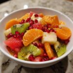 Close-up of a bright winter fruit salad featuring kiwi, orange segments, watermelon, and pomegranate seeds in a white bowl.