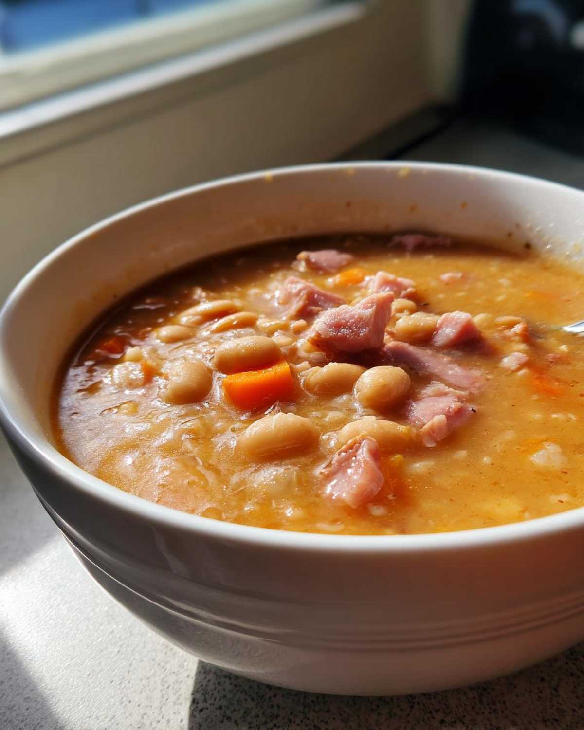 Close-up of a white bowl filled with thick, comforting navy bean soup featuring white beans, diced carrots, and pieces of ham.