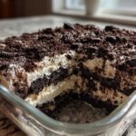 A close-up view of a rich, layered dirt cake featuring dark chocolate cookie crumbs and creamy filling in a clear glass baking dish.