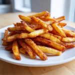 A close-up of a large pile of golden, seasoned air fryer french fries stacked on a white plate.