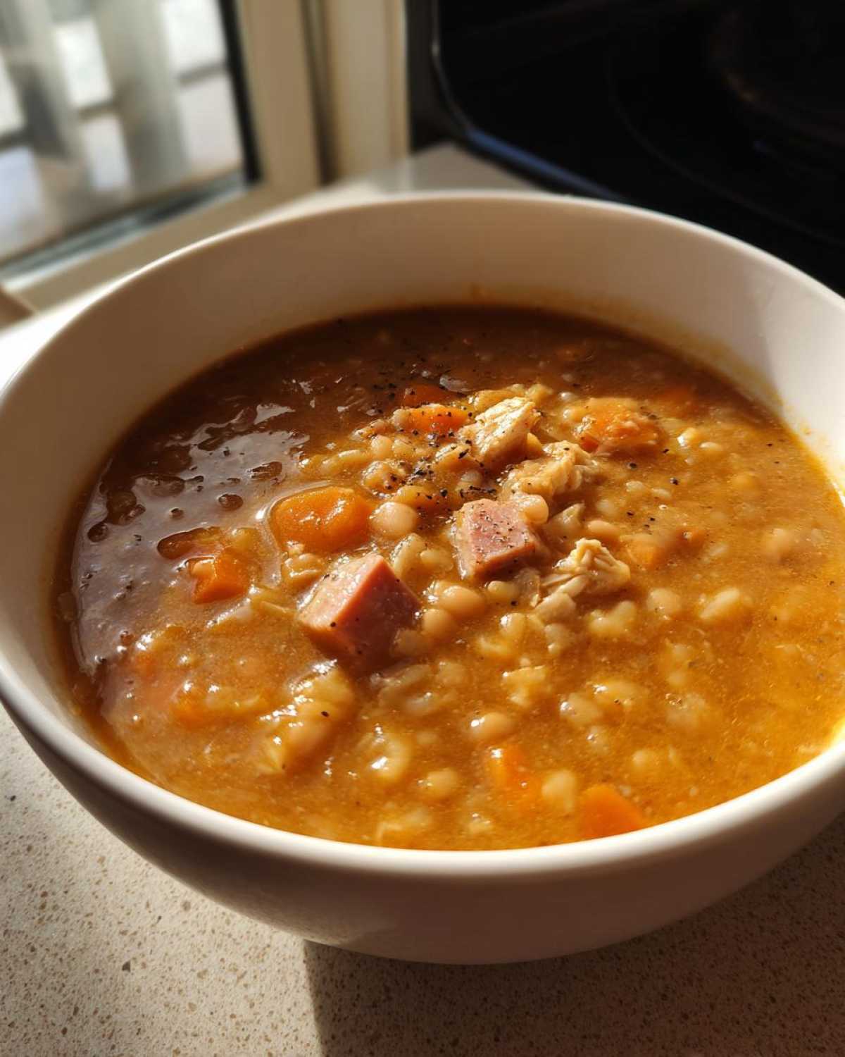A close-up of a white bowl filled with thick, hearty navy bean soup, featuring beans, carrots, ham, and pepper.