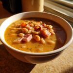 Close-up of a creamy, orange-hued bowl of navy bean soup loaded with ham chunks and vegetables.
