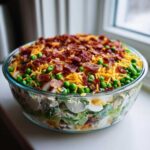 A large glass bowl filled with the amazing 7 layer salad, topped generously with peas, shredded cheddar cheese, and crispy bacon bits.