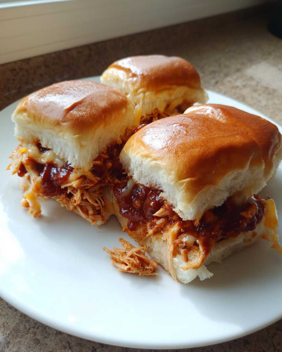 Close-up of three freshly made bbq chicken sliders filled with shredded chicken, BBQ sauce, and melted cheese on a white plate.