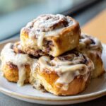 A close-up stack of soft, freshly baked cinnamon buns generously topped with white cream cheese frosting.