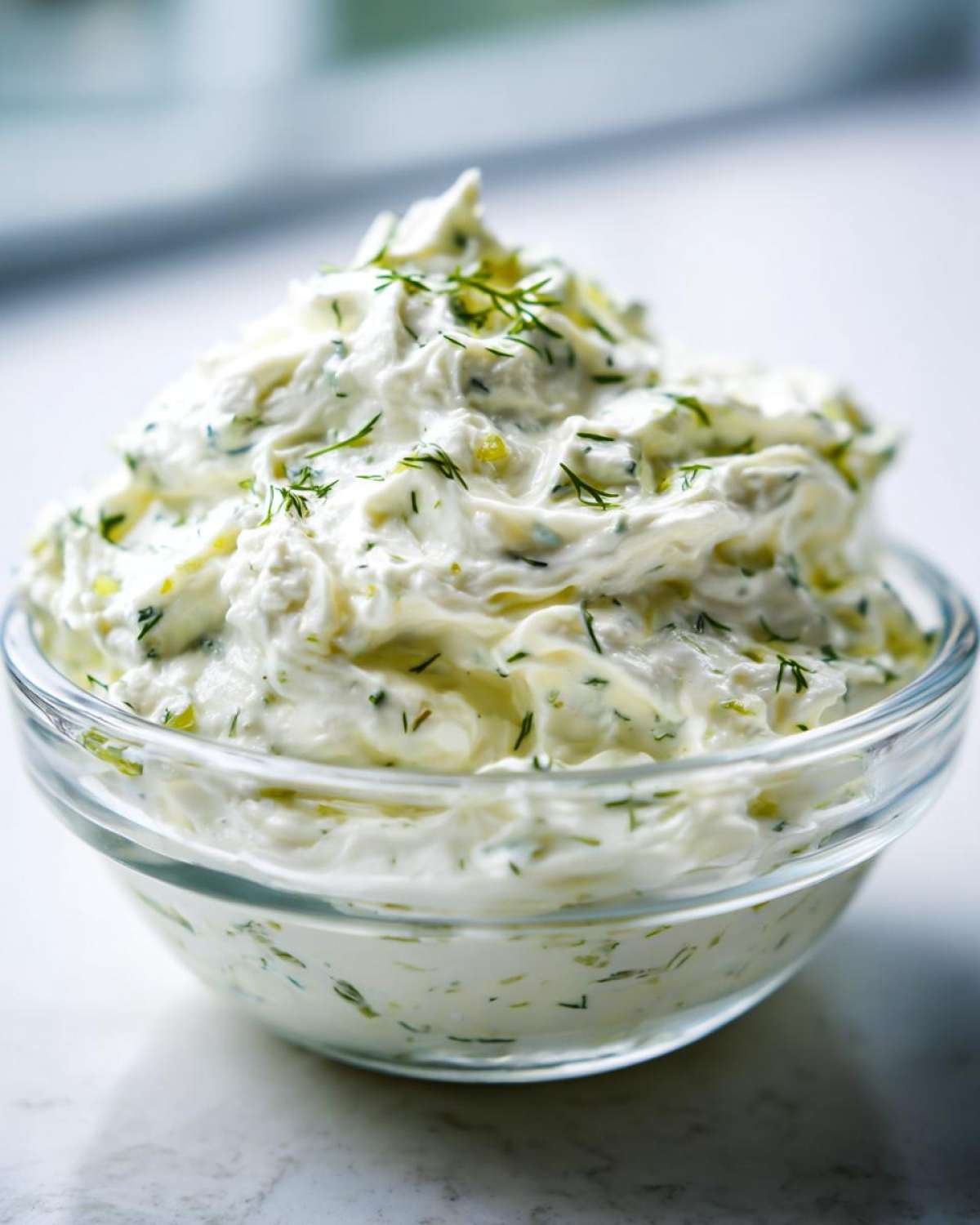 Close-up of a thick, creamy homemade pickle dip topped with fresh dill in a clear glass bowl.