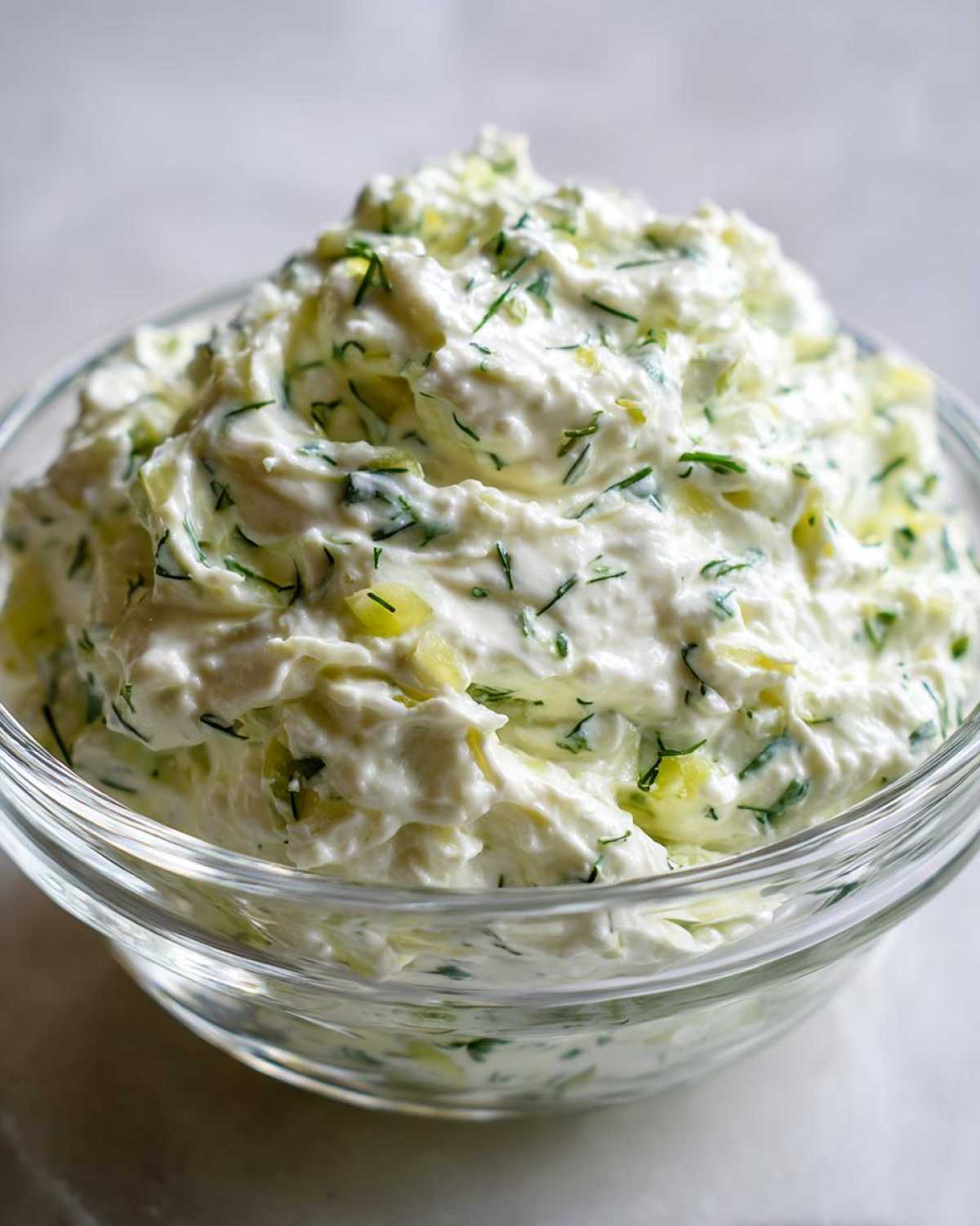 Close-up of creamy, white pickle dip mixed with chopped dill and pickle pieces in a clear glass bowl.