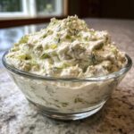 A close-up of a mound of creamy, chunky pickle dip, flecked with dill and pickle pieces, served in a clear glass bowl.