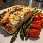 A close-up of a fillet of Baked Boursin Salmon topped with melted cheese, served with roasted asparagus and cherry tomatoes.