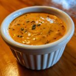 Close-up of a vibrant, orange-hued remoulade sauce speckled with herbs and spices, served in a white fluted ramekin.