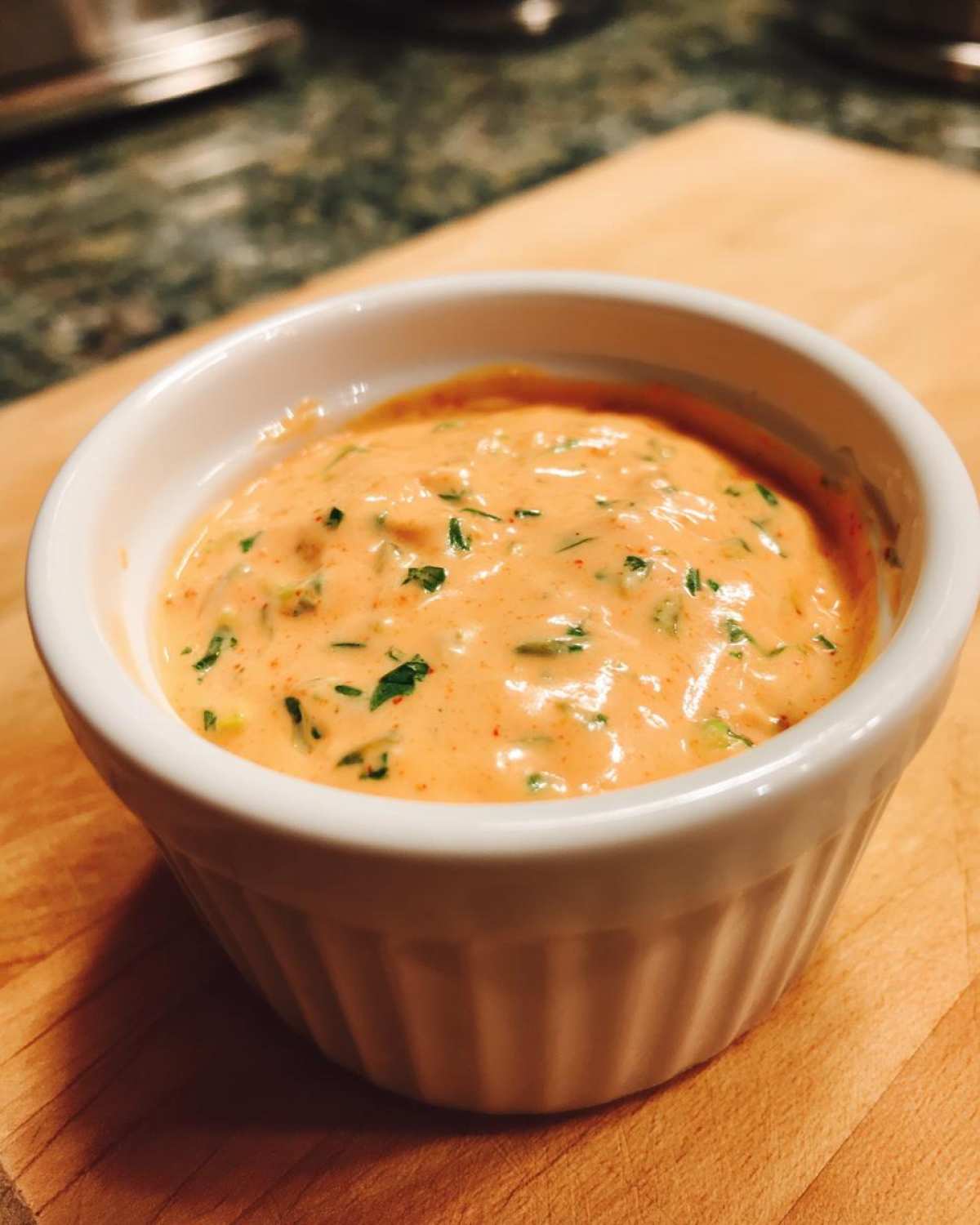 A close-up view of a small white ramekin filled with creamy, orange-hued remoulade sauce speckled with green herbs.