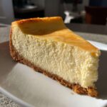 A close-up, sunlit slice of creamy baked cheesecake recipe on a white plate.