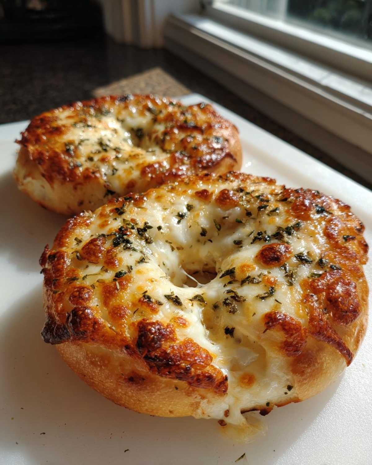 Close-up of two golden brown pizza bagels topped with melted, bubbly cheese and herbs, ready to eat.