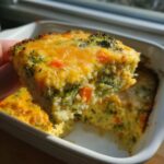 A hand lifting a square slice of baked Cheesy Veggie Quinoa Casserole topped with melted cheddar cheese.