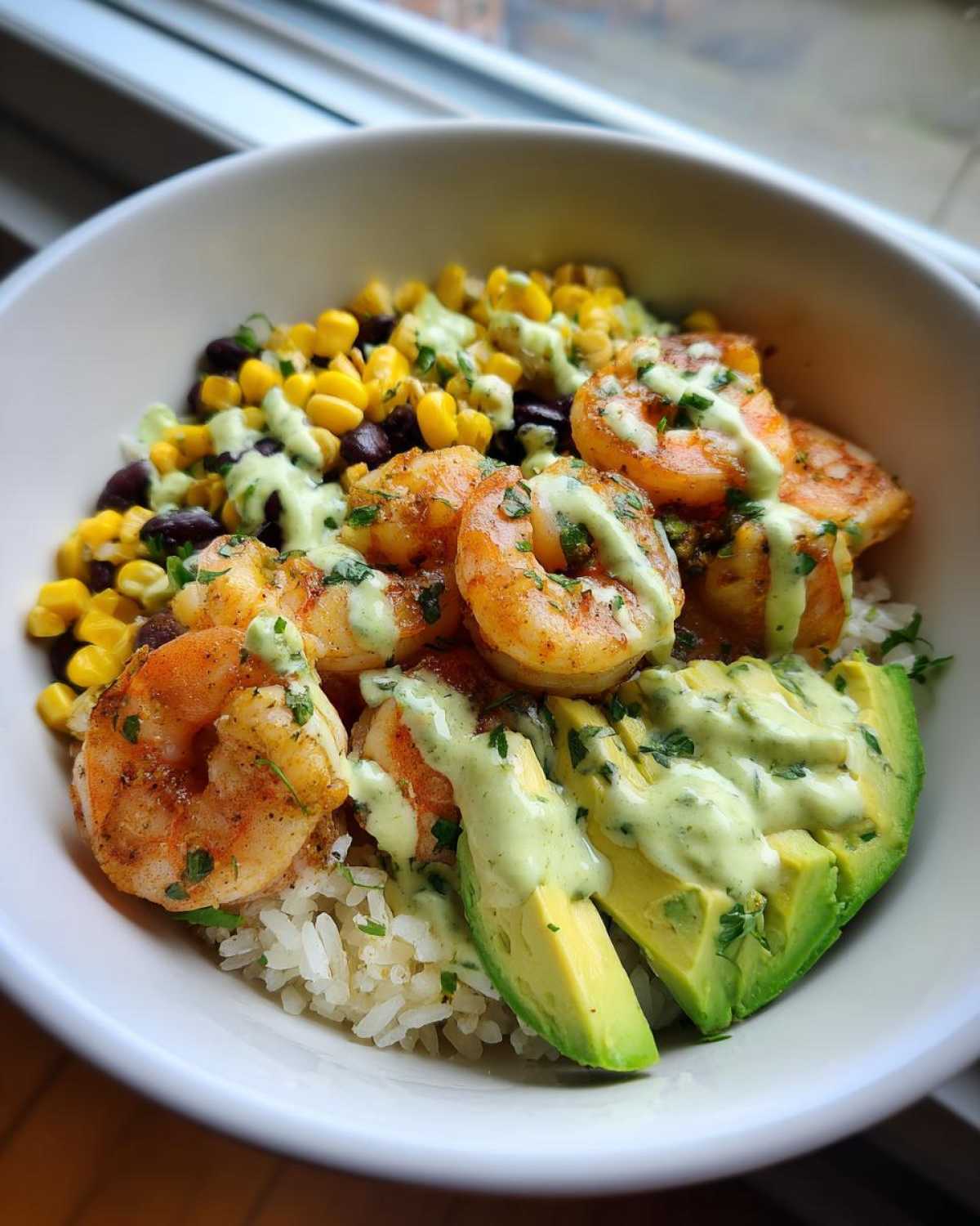 A close-up of a Chipotle Lime Shrimp Bowl featuring seasoned shrimp, rice, corn, black beans, avocado slices, and a creamy green drizzle.