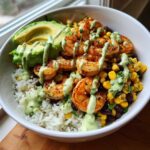 A close-up of a Chipotle Lime Shrimp Bowl featuring seasoned shrimp, white rice, black beans, corn, avocado slices, and a creamy green drizzle.