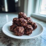 A stack of homemade chocolate oat protein balls resting on a small white plate near a window.