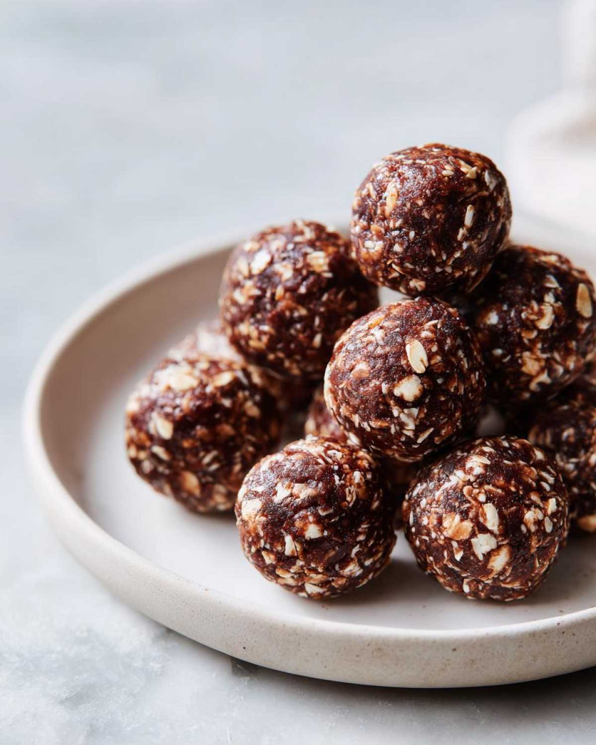 A close-up stack of dark chocolate protein balls mixed with visible rolled oats, served on a light ceramic plate.