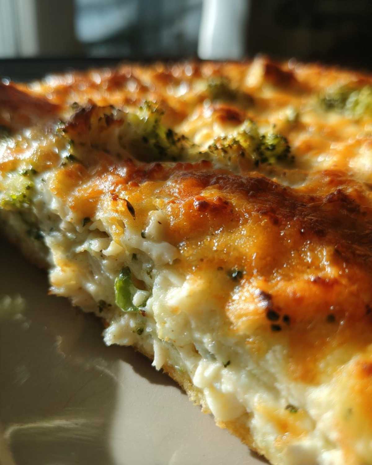 A close-up, sunlit shot of a slice of Low Carb Chicken Casserole showing creamy filling, chicken, and broccoli under a bubbly, golden-brown cheese crust.
