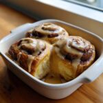 Close-up of three freshly baked Cookie Butter Cinnamon Rolls generously topped with a thick, dripping cookie butter glaze in a white baking dish.