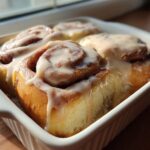 Close-up of soft, golden Cookie Butter Cinnamon Rolls topped with thick, creamy white glaze in a white baking dish.
