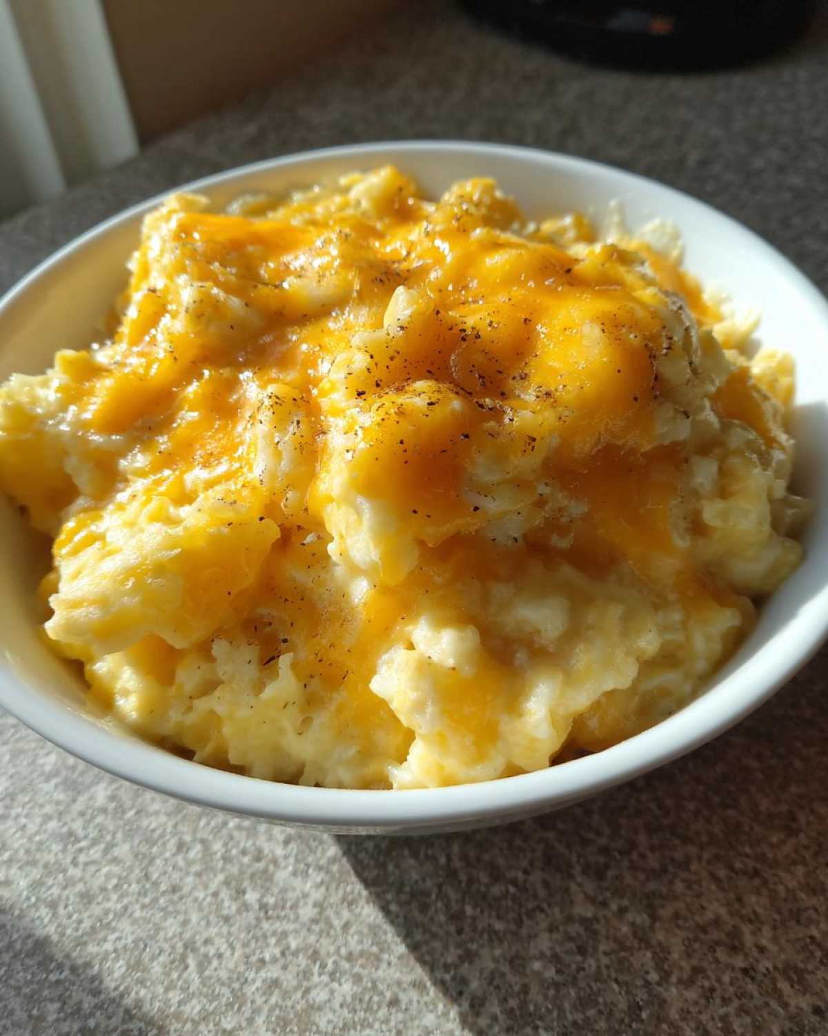 A close-up of a white bowl filled with creamy crockpot cheesy potatoes topped with melted cheddar cheese and black pepper.