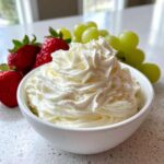 A white bowl filled with fluffy, creamy fruit dip, served alongside fresh strawberries and green grapes.